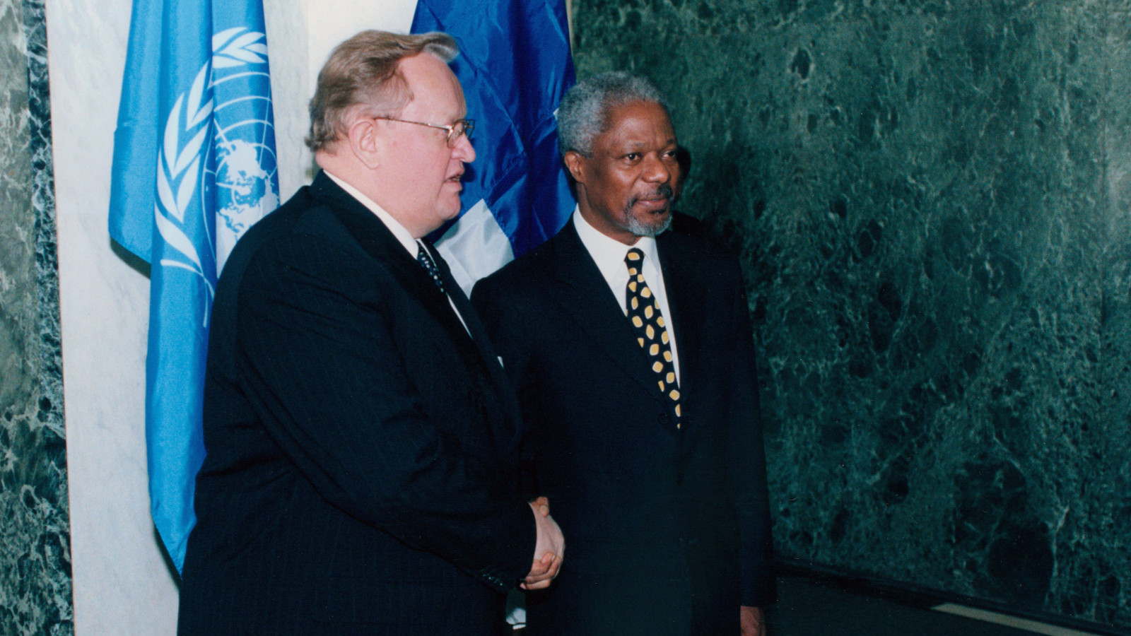 Ahtisaari and Kofi Annan shaking hands in front of flags of UN and Finland.